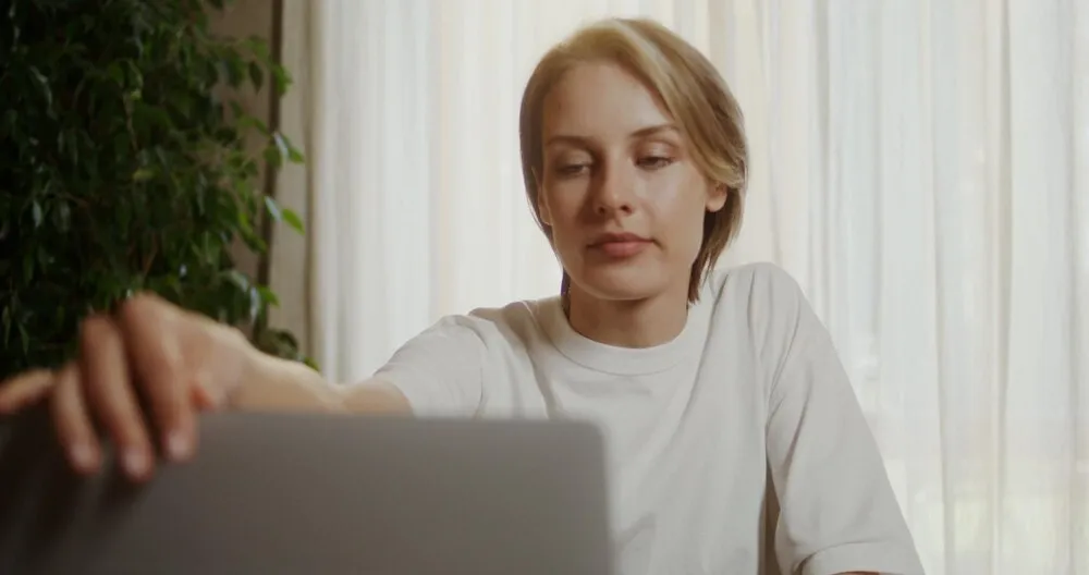 Młoda kobieta siada przy stole śniadaniowym, otwiera laptopa i pije kawę.
