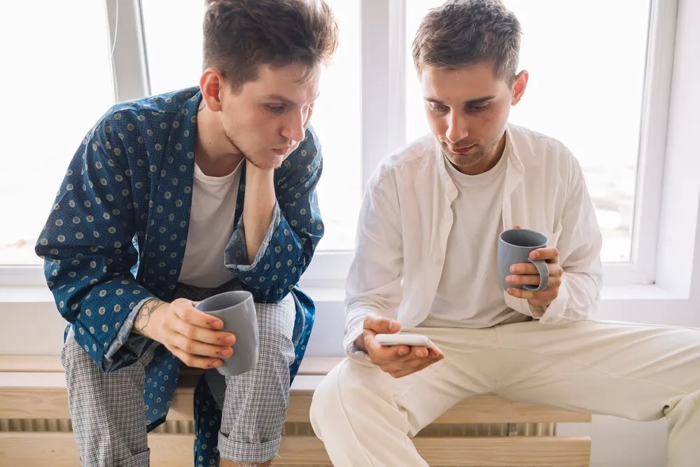 przystojni mężczyźni patrzący w telefon komórkowy i trzymający filiżankę kawy w dłoni